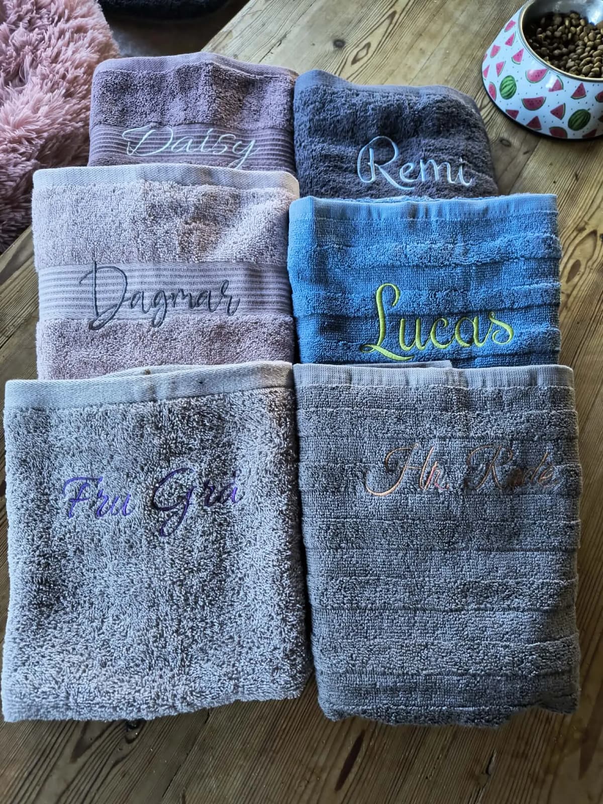 Seks foldede håndklæder i forskellige farver med broderede navne som Daisy, Remi, Dagmar, Lucas og Fru Grå